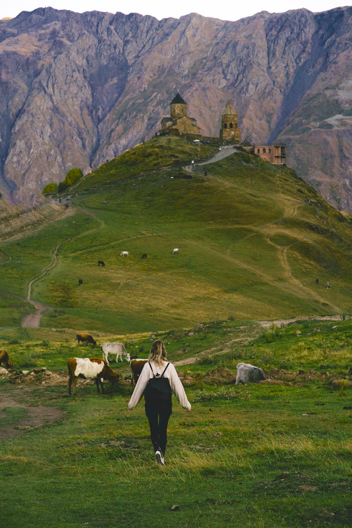 A woman walks on a grassy path toward Gergeti Trinity Church, surrounded by the stunning Caucasus Mountains.